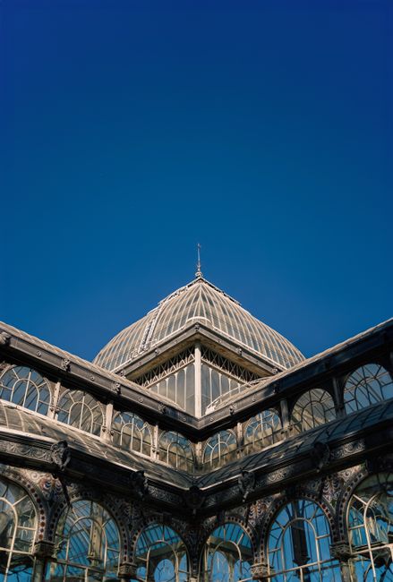A close-up, upward-facing view of a historic glass and iron conservatory or greenhouse structure, with ornate arched windows and intricate metalwork framing the glass panels. The building features a prominent, domed roof with decorative detailing, set against a clear, deep blue sky. The structure appears to be part of a larger architectural complex, with visible terraces and leaded window sections. The lighting highlights the reflective surfaces of the glass and the textured finishes of the metal framework, emphasizing the detailed craftsmanship. While the image focuses on the architectural design, it subtly suggests the importance of maintaining or clearing surrounding areas if the site were used for events or exhibitions, which could be supported by professional waste management services such as those provided by waste clearance specialists. The scene captures the historic elegance of the building, aligning with the context of private waste management or alternative collection methods often associated with unique or heritage sites.