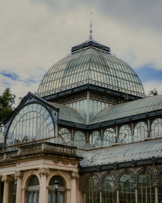 The image displays the intricate glass and metal roof structure of a historic greenhouse or conservatory, featuring large curved glass panels supported by metal frames, topped with a small ornamental spire. The greenhouse has a classical architectural base with ornate cornices, decorative columns, and detailed stonework, painted in light shades that contrast with the darker metalwork of the roofing. Surrounding leafy trees are partially visible against a partly cloudy sky, with sunlight reflecting off the glass surfaces, highlighting the transparency and texture of the glass panes. This structure exemplifies traditional on-site greenhouse design, combining aesthetic architectural elements with functional glass roofing, and it may serve as a backdrop for various horticultural or conservatory activities. Occasionally, waste clearance services like those from wasteclearancecrystalpalace.co.uk are needed to manage debris from building renovations or maintenance involving such historic structures, supporting private disposal or alternative waste handling outside of local authority waste collections.