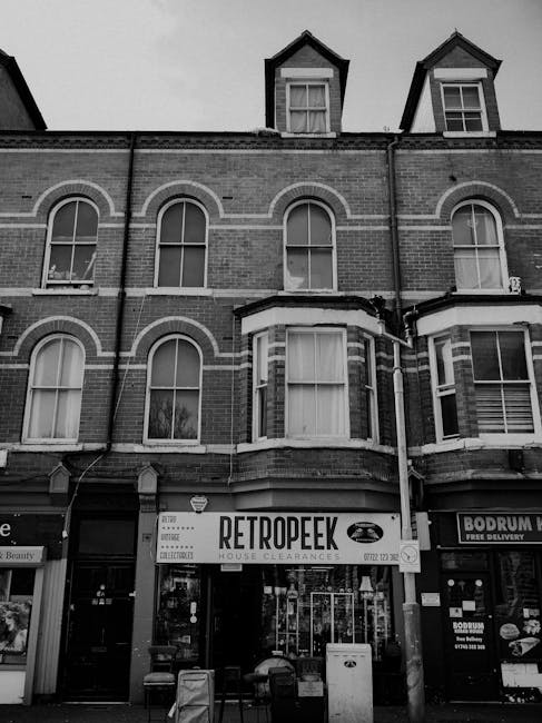 The image depicts the facade of a three-story brick building with a retail shop on the ground floor. The building has a traditional architectural style with arched window frames on the upper floors and a triangular gable roof with dormer windows. The shop's signage displays the company name 'RETROPEEK' along with the services offered, such as house clearances, indicating a focus on waste and rubbish removal. In front of the shop, there are several items, possibly rubbish or materials awaiting collection, including what appears to be a small container or bin. The surrounding environment includes neighboring shopfronts, one of which appears to be a food delivery service, and a sidewalk with a few outdoor chairs and miscellaneous objects. The scene is captured in black and white, emphasizing the commercial setting and the role of independent waste disposal services like those provided by wasteclearancecrystalpalace.co.uk in managing rubbish accumulation, particularly in urban residential or retail areas.