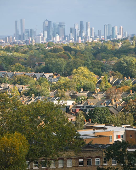 A panoramic view of a suburban area with rows of houses featuring pitched roofs and chimneys, situated among a dense canopy of green trees with various shades of foliage, some turning yellow and orange, indicating autumn. In the background, a modern city skyline with tall skyscrapers and glass facades rises against a partly cloudy sky, creating a stark contrast between the residential and urban environments. The scene is illuminated by natural daylight, with shadows cast by the trees and buildings, and appears to be taken from an elevated vantage point, possibly a hill or taller building, emphasizing the separation between the quiet, leafy suburb and the bustling city skyline beyond. The image highlights a peaceful residential zone adjacent to a vibrant urban centre, reflecting the proximity of private housing to opportunities for alternative waste collection and rubbish removal services, which are often utilized in such areas for efficient clearance of household waste by companies like wasteclearancecrystalpalace.co.uk.