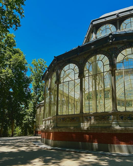 A detailed view of an ornate, vintage glass conservatory situated outdoors, featuring large arched windows with intricate metal framework and decorative elements. The structure's exterior showcases a combination of glass panels and carved stone or cast iron supports, with a curved roof design and decorative detailing along the sill and upper sections. The conservatory is positioned on a paved area, with lush green trees surrounding it, casting dappled shadows on the ground. The sky above is bright blue with no visible clouds, indicating a clear, sunny day. This scene provides context for a private or historic garden setting where independent waste clearance or rubbish removal services might be employed to maintain the area’s aesthetic, aligning with the theme of onsite or alternative waste handling that avoids reliance on local authority collections. Waste clearance specialists, like those at wasteclearancecrystalpalace.co.uk, could be involved in clearing debris or waste generated during maintenance or renovation activities around such historic structures, ensuring a clean environment without disrupting public access or preserving the site’s visual integrity.