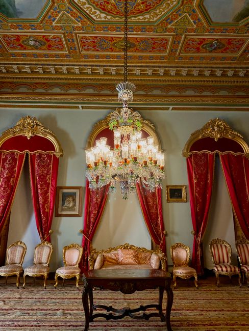 The image depicts an opulent interior room featuring a large, ornate crystal chandelier hanging from a richly decorated ceiling with intricate, colorful patterns. The walls are painted in a light shade, complemented by elegant draped red velvet curtains with gold trim and tassels, framing tall windows or alcoves. In front of the curtains, there is a classic wooden table with a dark finish, surrounded by six upholstered armchairs and a matching sofa, all with light-colored fabric and carved wooden frames, arranged in a semi-symmetric formation. The room's décor includes small framed paintings hung on the walls, adding to the luxurious ambiance. The richly patterned carpet on the floor enhances the regal atmosphere, while the overall scene exudes historical grandeur and classical elegance, typical of a stately mansion or palace interior. This setting may serve as a backdrop for formal gatherings, and while not directly related to rubbish removal, the image emphasizes the importance of maintaining pristine, elegant spaces, which companies like wasteclearancecrystalpalace.co.uk help preserve by offering alternative waste handling solutions.