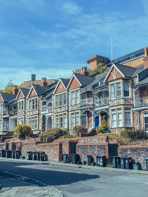 A row of Victorian-style terraced houses with ornate white decorative trim, bay windows, and small front gardens bordered by brick walls. The houses are situated on a sloped street with black wheelie bins lined up along the pavement in the foreground. Bright daylight illuminates the scene, with a clear blue sky overhead. In the background, modern commercial or office buildings are visible behind the houses. The image captures a typical residential street scenario where private waste collection, such as that provided by waste clearance services like [COMPANY_NAME], might be involved in on-site clearance or alternative rubbish removal for homeowners or property managers in the area.