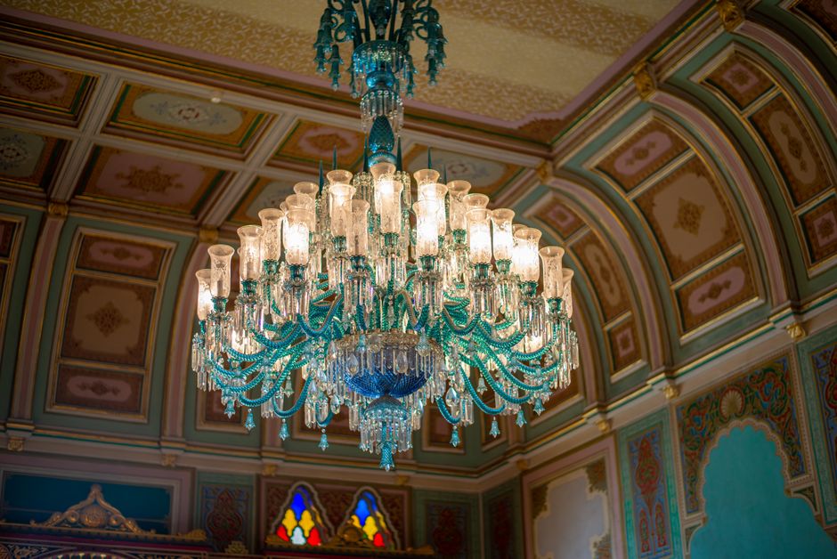 The image depicts an opulent interior room featuring a large, ornate crystal chandelier hanging from a richly decorated ceiling with intricate, colorful patterns. The walls are painted in a light shade, complemented by elegant draped red velvet curtains with gold trim and tassels, framing tall windows or alcoves. In front of the curtains, there is a classic wooden table with a dark finish, surrounded by six upholstered armchairs and a matching sofa, all with light-colored fabric and carved wooden frames, arranged in a semi-symmetric formation. The room's décor includes small framed paintings hung on the walls, adding to the luxurious ambiance. The richly patterned carpet on the floor enhances the regal atmosphere, while the overall scene exudes historical grandeur and classical elegance, typical of a stately mansion or palace interior. This setting may serve as a backdrop for formal gatherings, and while not directly related to rubbish removal, the image emphasizes the importance of maintaining pristine, elegant spaces, which companies like wasteclearancecrystalpalace.co.uk help preserve by offering alternative waste handling solutions.