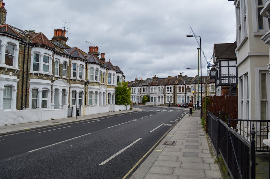A quiet residential street lined with Victorian-style terraced houses featuring ornate white facades, bay windows, and reddish-brown tiled roofs with chimneys. The buildings are positioned close to the pavement, which is edged by black metal railings on the right side and a wide grey sidewalk. The street appears to be recently paved with dark asphalt, with visible white road markings indicating parking bays. A single cyclist is seen riding along the road towards the distance, while a person is walking on the pavement on the right-hand side. Tall, modern streetlights are positioned intermittently along the sidewalk, and in the background, construction cranes are visible in the sky, suggesting ongoing development. The overcast sky casts diffuse, even lighting over the scene. This urban environment reflects a typical area where private waste clearance, such as rubbish removal services by companies like wasteclearancecrystalpalace.co.uk, might be involved in maintaining the cleanliness of residential streets.