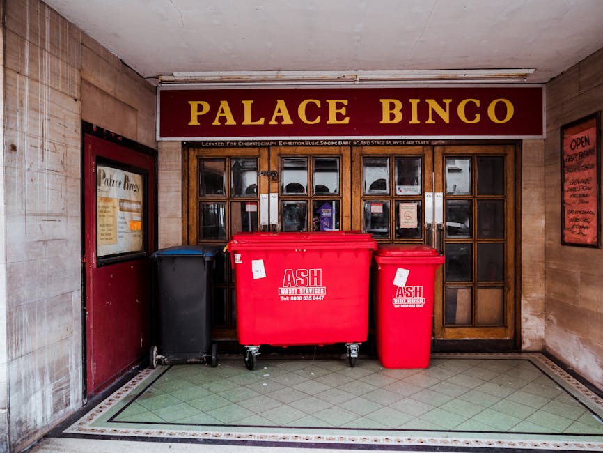 Inside a building entrance displaying the sign 'Palace Bingo' above wooden-framed glass doors with multiple small panes; in front of the doors, there are three large wheeled waste bins placed on a tiled floor with a green and beige pattern. The bins include a black bin on the left, a large red bin in the center, and a smaller red bin on the right, all labeled with waste collection company branding and contact information. The scene is lit by indoor lighting, with walls on either side that are partly tiled and partly painted, suggesting an indoor area possibly near a commercial or entertainment venue. The presence of waste collection bins indicates on-site waste management, consistent with private rubbish disposal services often used in commercial settings to handle refuse outside of local authority collection, fitting into the broader context of rubbish removal options for such premises.