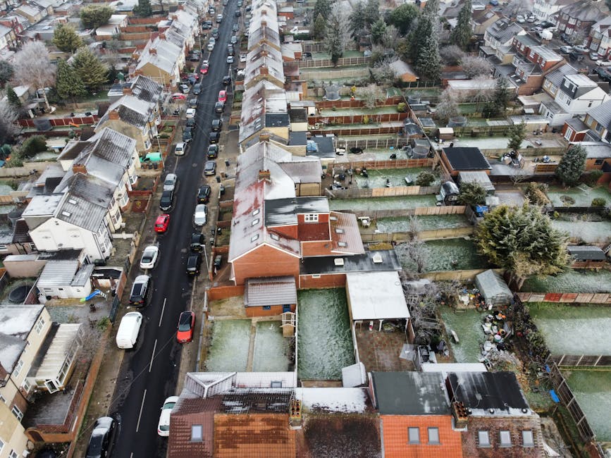 A quiet residential street lined with Victorian-style terraced houses featuring ornate white facades, bay windows, and reddish-brown tiled roofs with chimneys. The buildings are positioned close to the pavement, which is edged by black metal railings on the right side and a wide grey sidewalk. The street appears to be recently paved with dark asphalt, with visible white road markings indicating parking bays. A single cyclist is seen riding along the road towards the distance, while a person is walking on the pavement on the right-hand side. Tall, modern streetlights are positioned intermittently along the sidewalk, and in the background, construction cranes are visible in the sky, suggesting ongoing development. The overcast sky casts diffuse, even lighting over the scene. This urban environment reflects a typical area where private waste clearance, such as rubbish removal services by companies like wasteclearancecrystalpalace.co.uk, might be involved in maintaining the cleanliness of residential streets.